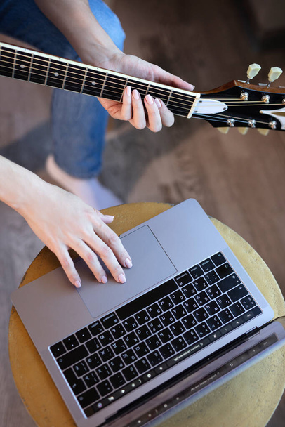 Young woman sits in a room on an armchair, learns to play guitar online. Online education, online music lessons, online vocal and guitar training. Stay home. - Φωτογραφία, εικόνα
