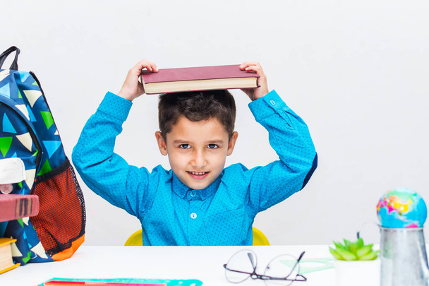 Koyu renk saçlı, mavi gömlekli küçük bir çocuk bir masada oturuyor ve kafasına bir kitap tutuyor. Anaokulundaki çocuklarda dikkat eksikliği.. - Fotoğraf, Görsel