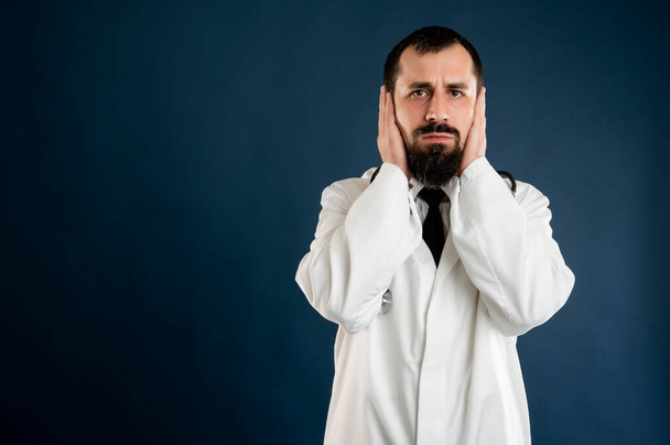 Portrait de médecin masculin avec stéthoscope en uniforme médical couvrant les oreilles comme un concept sourd posant sur un fond bleu isolé
. - Photo, image