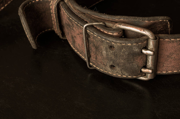 Cracked very old leather belt with a double metal buckle. Close-up of a wide dirty belt or collar on a dark table. Collectibles vintage clothing accessories. Selective focus. - Foto, Bild