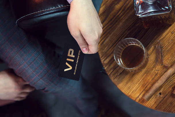Man holds VIP member card in his hand. View from the top on the gentleman's hand that holds exclusive VIP membership card next to the wooden table with whisky in carafe and glass with cigar. - 写真・画像