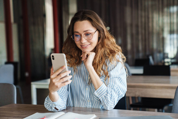 Image of joyful charming woman in eyeglasses smiling and using cellphone while studying at classroom - Foto, Bild
