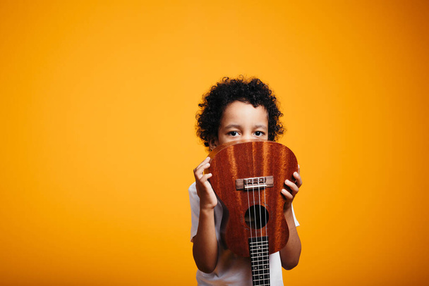 Curly african american boy in white t-shirt hiding behind ukulele guitar on orange background - Photo, Image