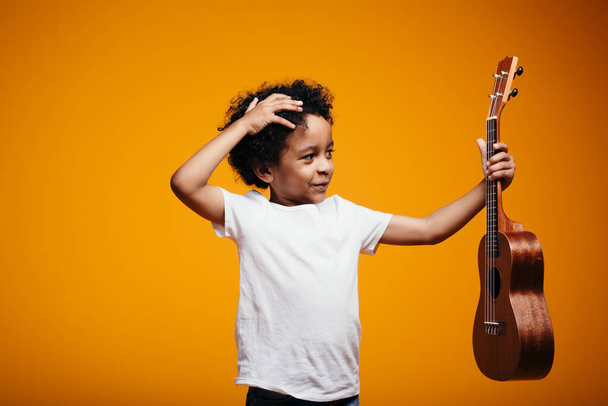 Curly-haired boy Maracan thoughtfully scratches his head and looks at a ukulele guitar not knowing how to play it in the studio - Фото, зображення