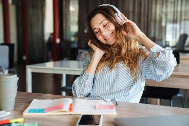 Image of happy young woman listening music with wireless headphones while studying at classroom - Fotografie, Obrázek
