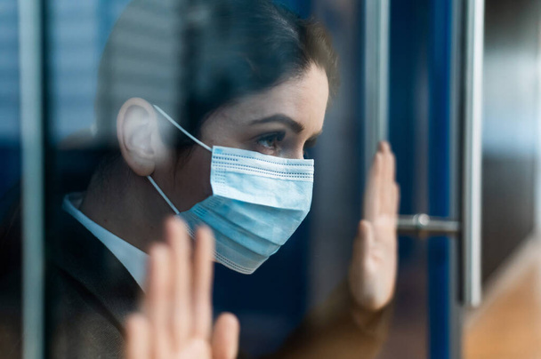 A sad quarantined woman in a business suit leaned against the window. Caucasian girl in a mask on isolation put her hands on a glass door. Maintaining social distance to prevent coronavirus. - Foto, afbeelding