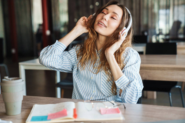Image of happy young woman listening music with wireless headphones while studying at classroom - 写真・画像
