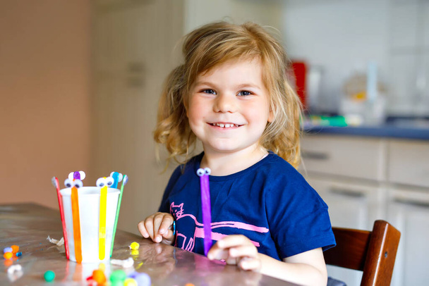 little toddler girl making craft lantern with paper cups, colorful pompoms and glue during pandemic coronavirus quarantine disease. Happy creative child, homeschooling and home daycare with parents - Photo, Image