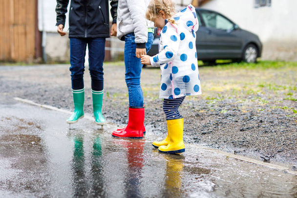 Κοντινό πλάνο τριών παιδιών, ενός μικρού κοριτσιού και δύο παιδιών αγοριών που φορούν κόκκινες, κίτρινες και πράσινες μπότες βροχής και περπατούν κατά τη διάρκεια του ύπνου. Χαρούμενα αδέρφια που πηδάνε σε λακκούβα. Διασκέδαση σε εξωτερικούς χώρους, ενεργό οικογένεια - Φωτογραφία, εικόνα