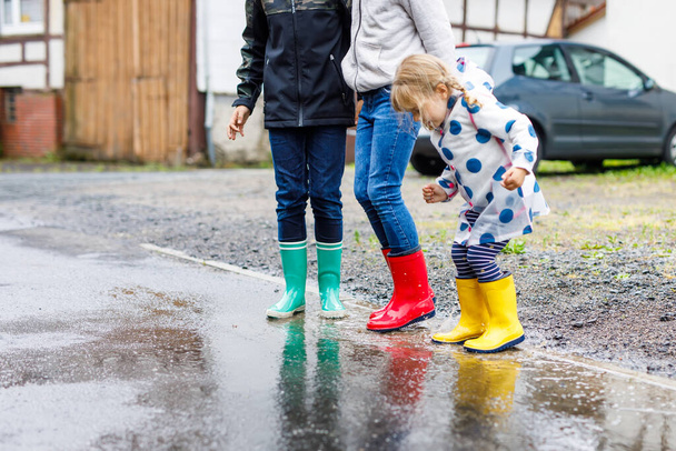 Közelkép három gyerekről, kislányról és két fiúról piros, sárga és zöld esőcsizmában, akik esőben sétálnak. Boldog testvérek ugranak a tócsába. Szórakozás szabadban, aktív család - Fotó, kép