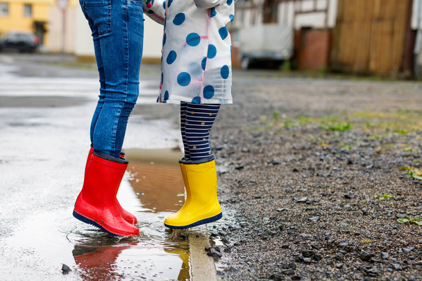 Großaufnahme von zwei Kindern, einem kleinen Mädchen und einem kleinen Jungen, die rote und gelbe Regenstiefel tragen und bei Schneeregen spazieren gehen. Glückliche Geschwister, Bruder und Schwester springen in die Pfütze. Spaß im Freien, aktive Familie - Foto, Bild