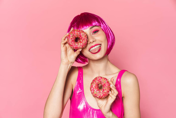 Image of young party girl wearing wig smiling and holding donuts isolated over pink background - Photo, Image