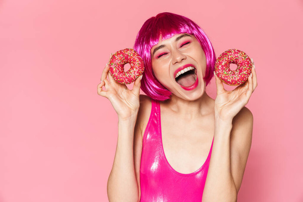 Image of young party girl wearing wig smiling and holding donuts isolated over pink background - Photo, image