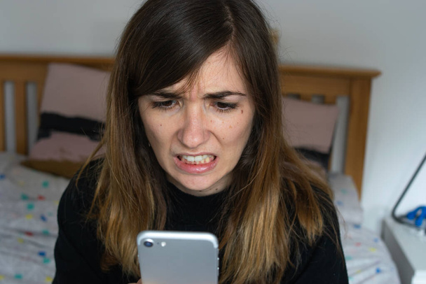 Zdjęcie młodej i atrakcyjnej kobiety z bardzo gniewnym wyrazem twarzy patrzącej na jej telefon. Złe wieści. - Zdjęcie, obraz