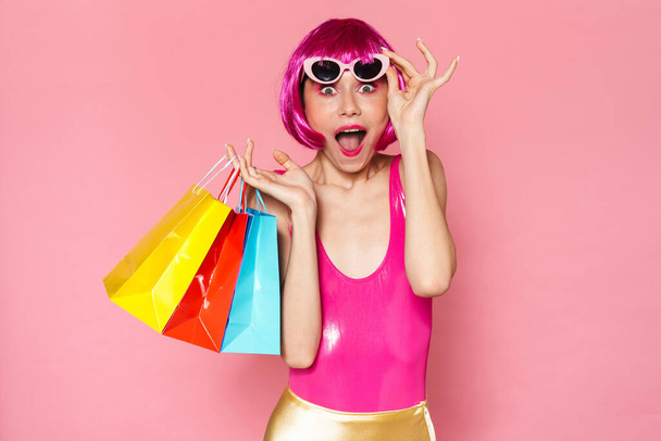 Image of excited girl wearing wig and sunglasses screaming and holding shopping bags isolated over pink background - Fotoğraf, Görsel