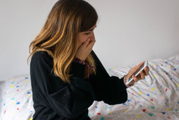 Zdjęcie młodej i atrakcyjnej dziewczyny z czarnym swetrem zakrywającym usta zmartwionym wyrazem twarzy podczas sprawdzania telefonu. Złe wieści. - Zdjęcie, obraz