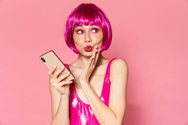 Image of young party girl wearing wig making kiss lips and using mobile phone isolated over pink background - Foto, Bild