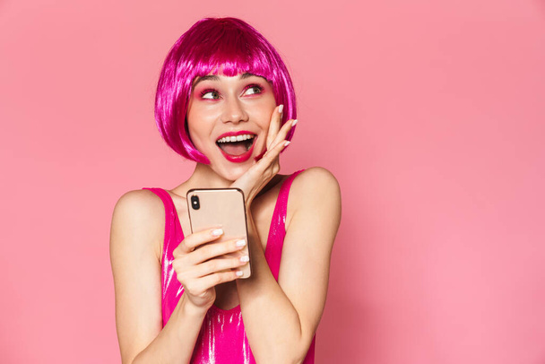 Image of young happy girl wearing wig smiling and holding cellphone isolated over pink background - Foto, immagini