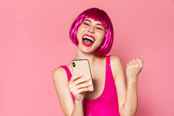 Image of young happy girl wearing wig smiling and holding cellphone isolated over pink background - Foto, Imagem