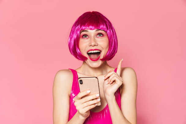 Image of young happy girl wearing wig smiling and holding cellphone isolated over pink background - Foto, Imagen