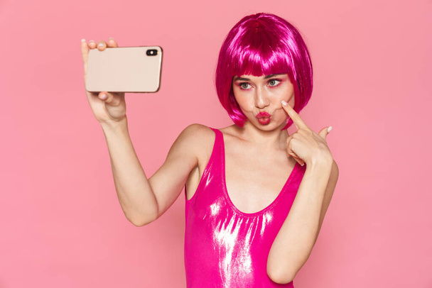 Image of young happy girl wearing wig smiling and taking selfie photo on cellphone isolated over pink background - Φωτογραφία, εικόνα