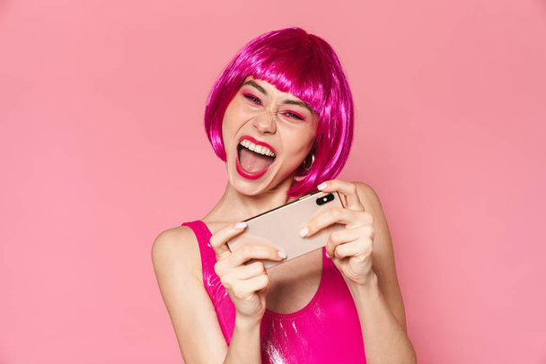 Image of young happy girl wearing wig smiling and playing video games on cellphone isolated over pink background - Φωτογραφία, εικόνα