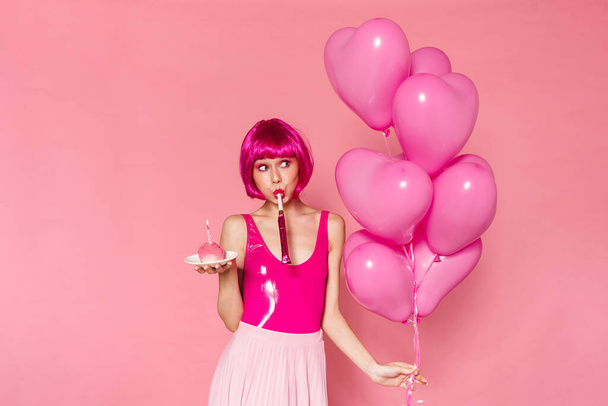 Image of amusing nice woman blowing in party horn while holding balloons and cake isolated over pink background - Zdjęcie, obraz