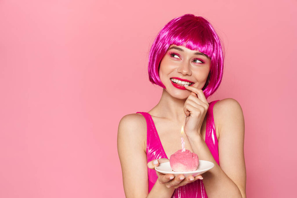 Image of excited cute woman in wig smiling and holding cake with candle isolated over pink background - Foto, immagini