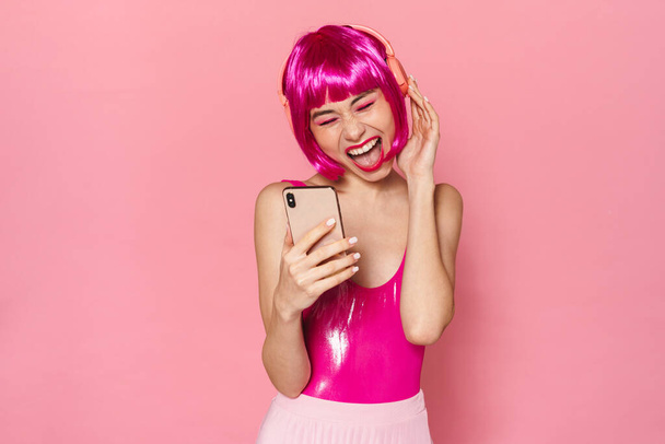 Image of excited pretty woman in wireless headphone singing and using cellphone isolated over pink background - Foto, imagen