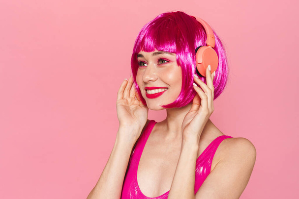 Kép vidám csinos nő parókában mosolyog, és használja a vezeték nélküli fejhallgató elszigetelt rózsaszín háttér - Fotó, kép