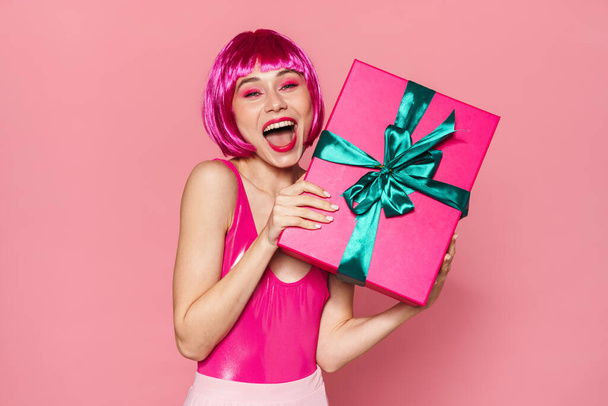 Image of happy woman in wig expressing surprise and holding gift box isolated over pink background - Fotó, kép
