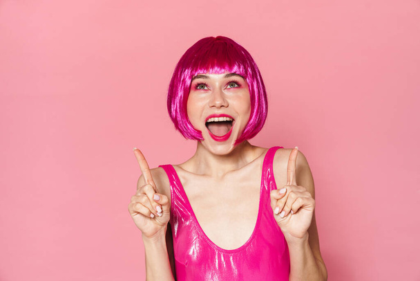 Image of excited girl wearing wig expressing surprise and pointing fingers upward isolated over pink background - 写真・画像