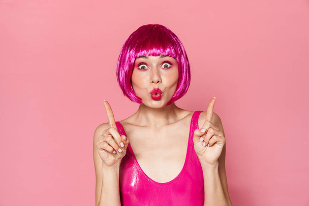 Image of amusing girl wearing wig making kiss lips and pointing fingers upward isolated over pink background - Foto, Bild