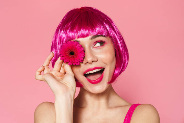 Imagem de mulher bonita alegre na peruca sorrindo enquanto posando com flor isolada sobre fundo rosa
 - Foto, Imagem
