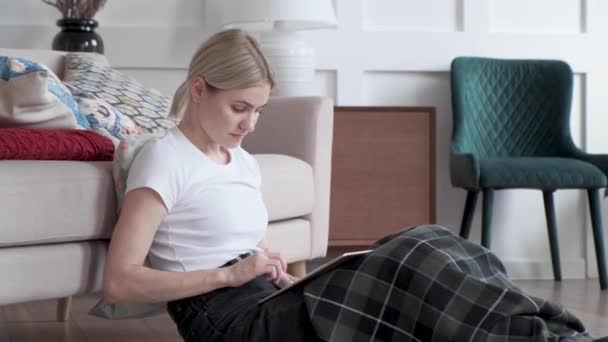 Mulher sorridente relaxante segurando tablet digital leitura e livro sentar perto de sofá em casa, senhora adulta sênior gostando de usar aplicativos almofada de computador navegando internet compras no sofá na sala de estar
 - Filmagem, Vídeo