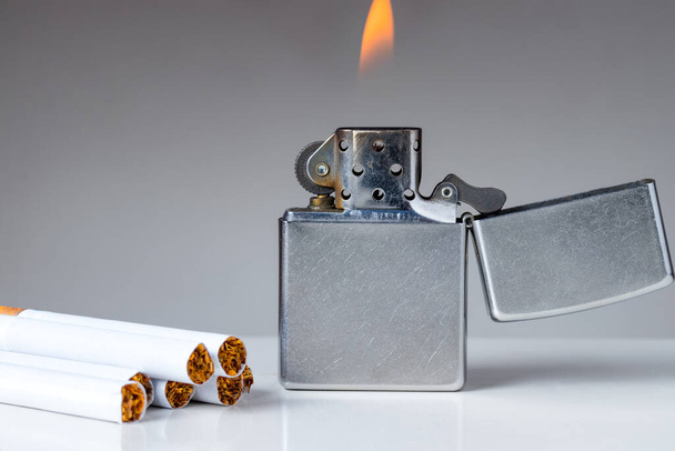 Zigaretten und Feuerzeug mit Flamme auf weißem und grauem Hintergrund. Nikotin- und Tabaksucht abstraktes Konzept. Kopierraum links. - Foto, Bild