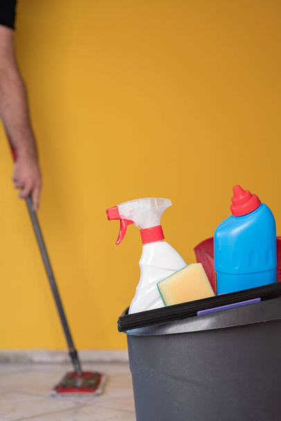 House spring cleaning items or supplies in a bucket. Caucasian man washing tile floor with a mop in a background. Vertical orientation. - Photo, Image