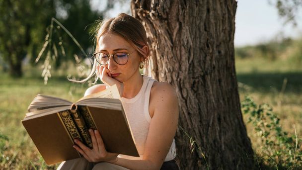 Λευκό ξανθό κορίτσι σκέφτεται διαβάζοντας ένα βιβλίο κάθεται κάτω από ένα δέντρο στο πάρκο περιτριγυρισμένο από τη φύση - Φωτογραφία, εικόνα
