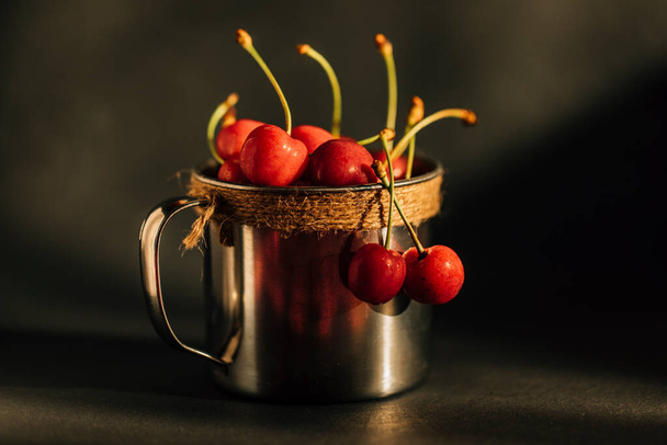 Wet organic cherries in vintage metal cup on wooden table. Selective focus. Dark rustic background with cherry and drops of water. - Fotó, kép