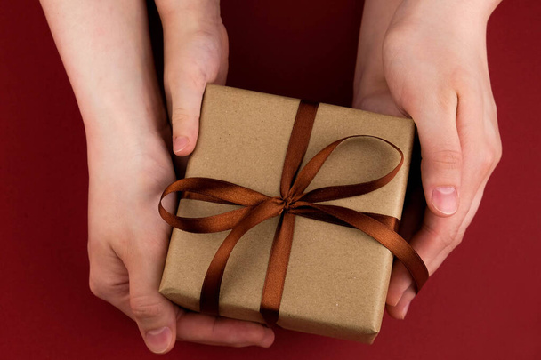 Female hands hold a gift box wrapped in craft paper and tied with a brown ribbon on a red background close-up - Fotoğraf, Görsel
