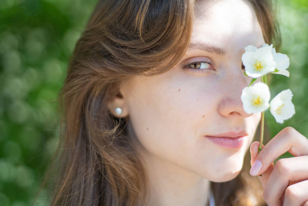 Portrait of a young woman close-up, covering her eye with a branch of jasmine. Beautiful smiling girl with flower in her hand - Fotoğraf, Görsel