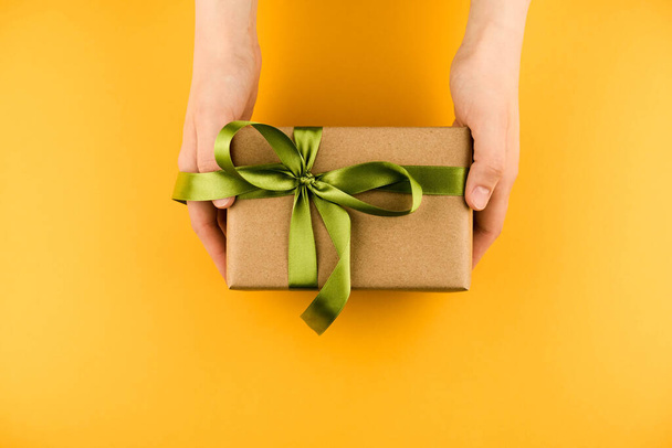Female hands hold a gift box wrapped in craft paper and tied with a green ribbon on a yellow background close-up - Foto, imagen