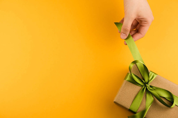 Female hands hold a gift box wrapped in craft paper and tied with a green ribbon on a yellow background close-up. Copy space. - Фото, зображення