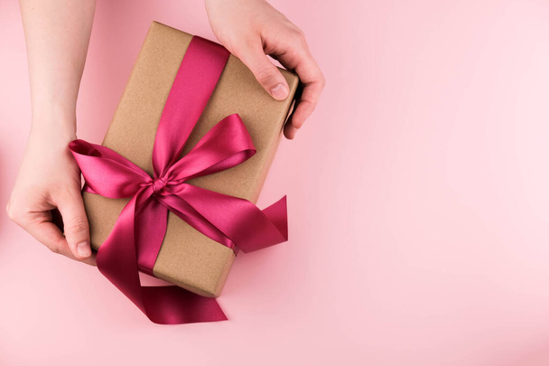 Female hands hold a gift box wrapped in craft paper and tied with a pink ribbon on a pink background close-up. Copy space. - Fotografie, Obrázek