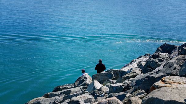 Mężczyźni łowiący ryby z kamiennego muru w porcie do błękitnego oceanu. - Zdjęcie, obraz