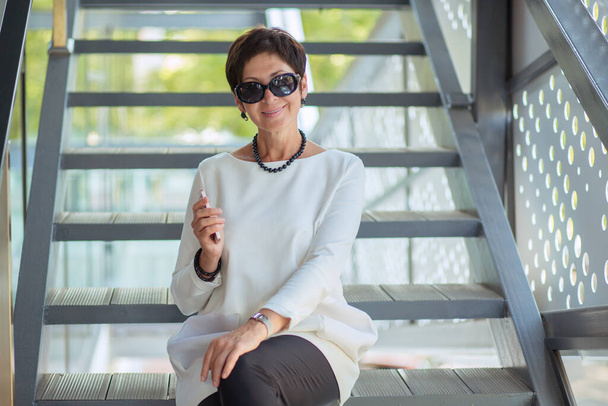 Cheerful middle-aged stylish woman in black sunglasses sitting on stairs on street relaxing looking at you camera and smiling happily wearing white blouse dress holding mobile in hand - Photo, Image