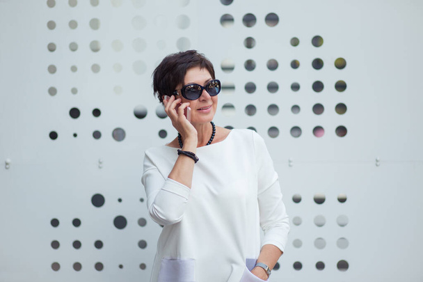 Trendy middle-aged woman wearing sunglasses and white dress speaking on smartphone cheerfully standing on street on a white metallic wall background - Foto, afbeelding