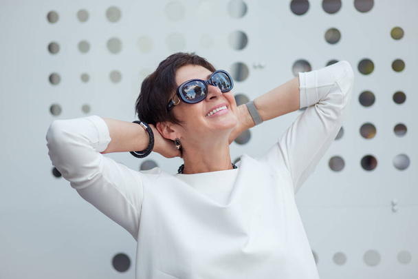 Smiling mature 50 years old aged woman day dreaming about summer leisure vacation in sunglasses hands behind head while standing near white metallic wall. Happy lady relaxing Pleasant memories concept - Photo, Image