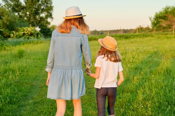 Rückansicht, zwei Mädchen gehen Händchen haltend auf einer grünen Sommerwiese - Foto, Bild