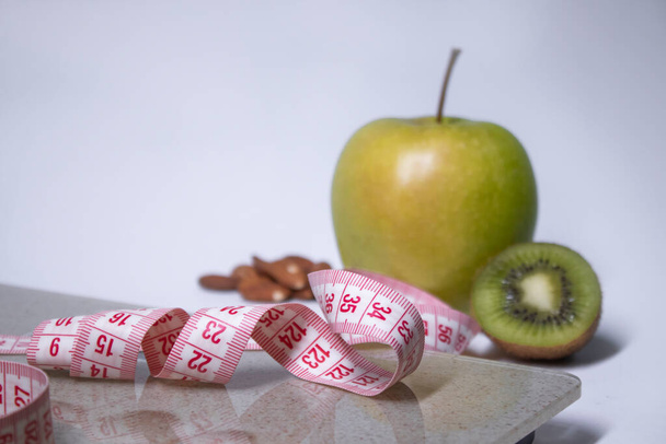 Red measuring tape, apple, kiwi and almonds near bathroom scales. Diet and Healthy life, loss weight, sport concept. Copy space. Isolated. White background - Photo, Image
